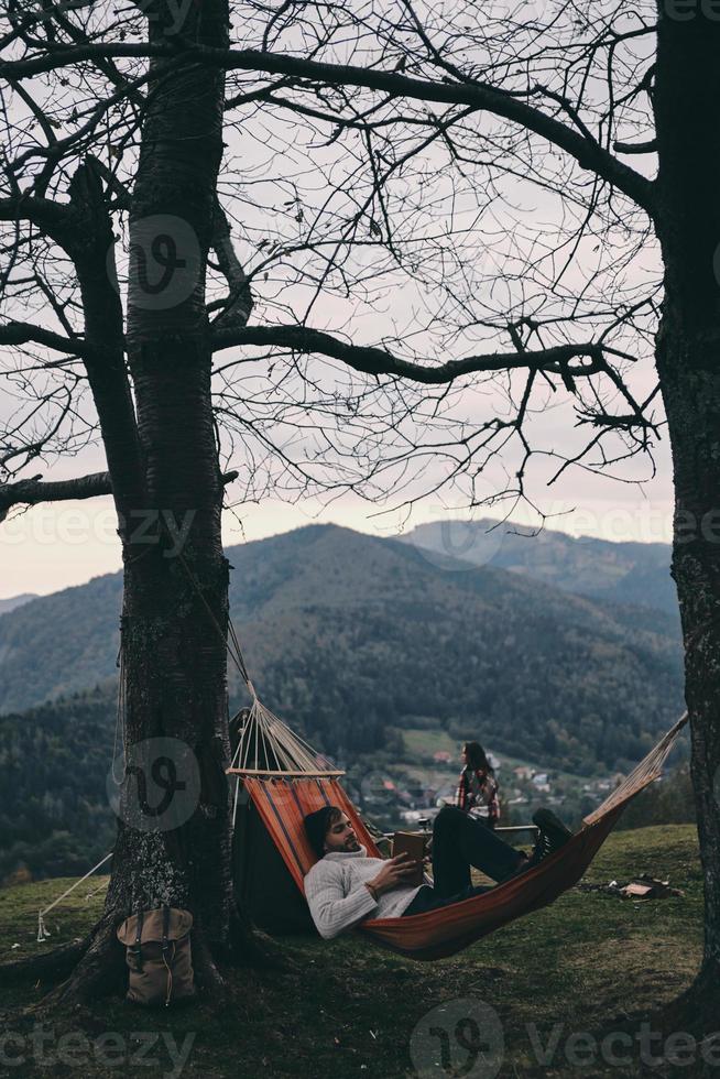 mejor que cualquier spa. apuesto joven acostado en una hamaca y leyendo un libro mientras acampa con su novia foto