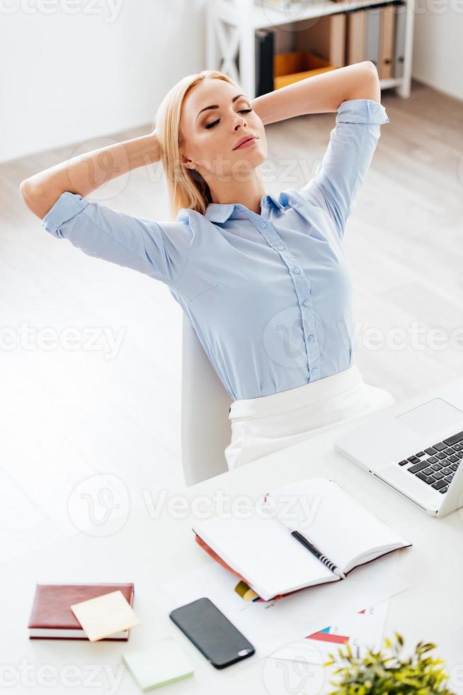 Little rest. Top view of beautiful young woman holding hands behind her head and keeping eyes closed while sitting at her working place photo
