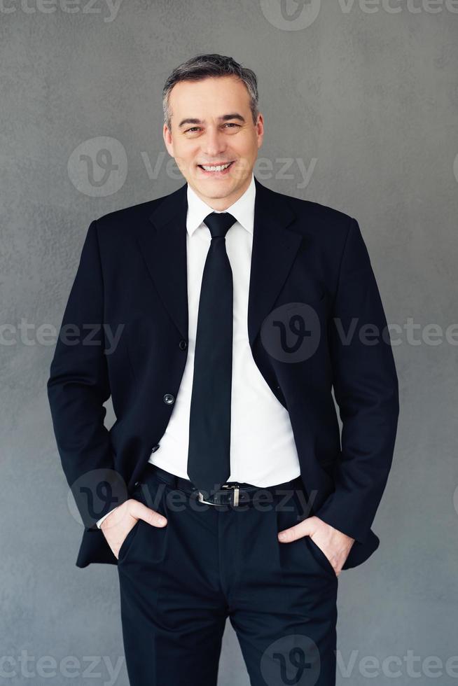 Professional and successful. Mature businessman keeping hands in pockets and looking at camera with smile while standing against grey background photo