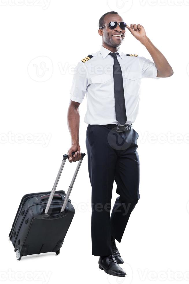 listo para un nuevo vuelo. feliz piloto africano en uniforme caminando y llevando maleta mientras está aislado en fondo blanco foto