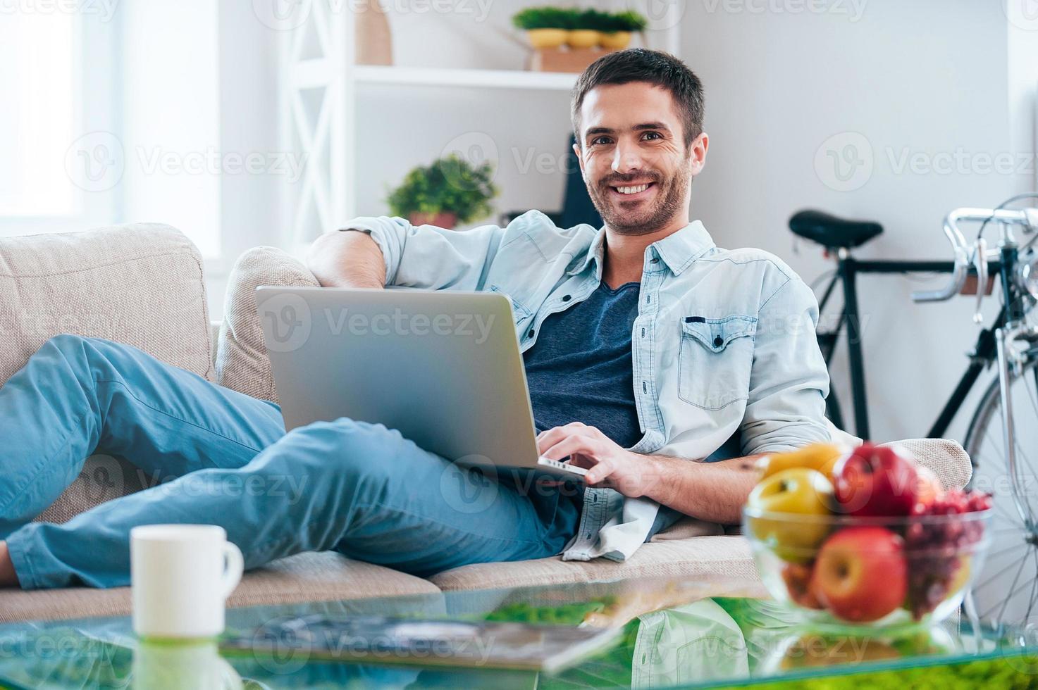 Carefree time at home. Handsome young man working on laptop and smiling while lying on the couch at home photo