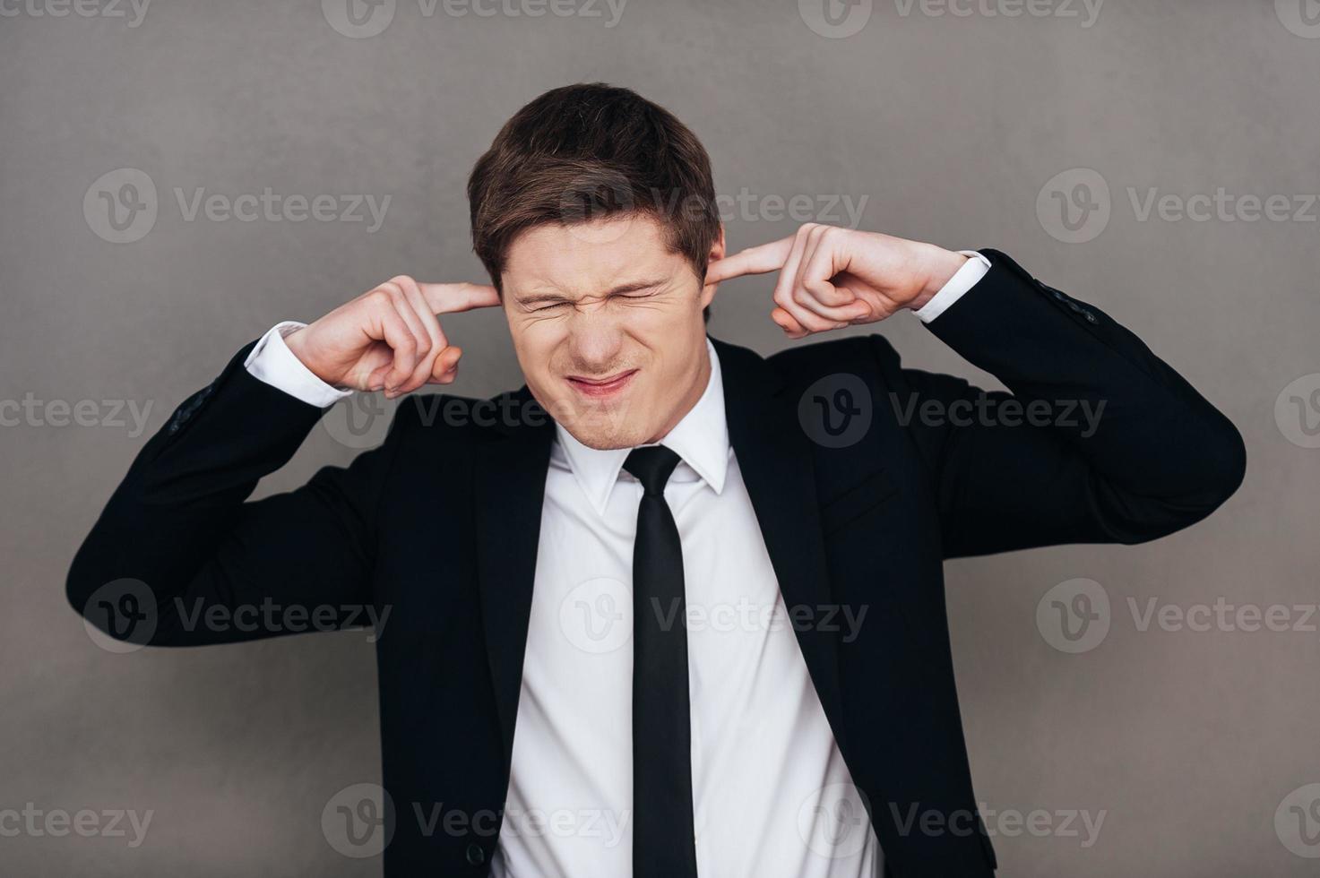 This sound is too loud. Frustrated young man in formalwear closing his ears with fingers and making a face while standing against grey background photo