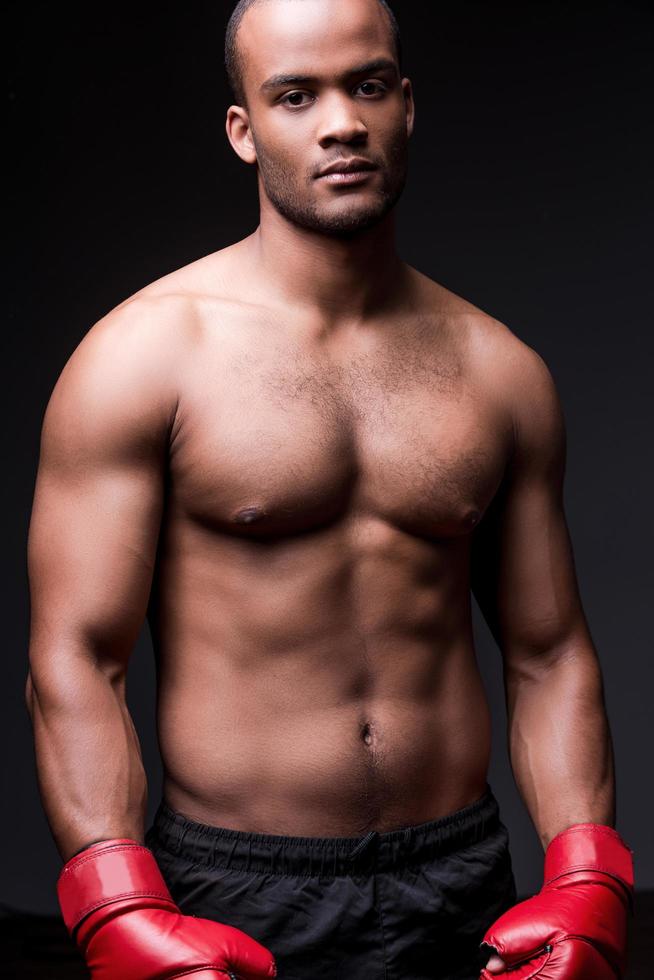 Energy inside him. Young shirtless African man in boxing gloves looking at camera while standing against grey background photo