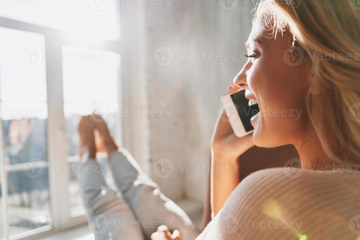 Rear view of beautiful young woman talking on her smart phone and smiling while sitting near the window at home photo