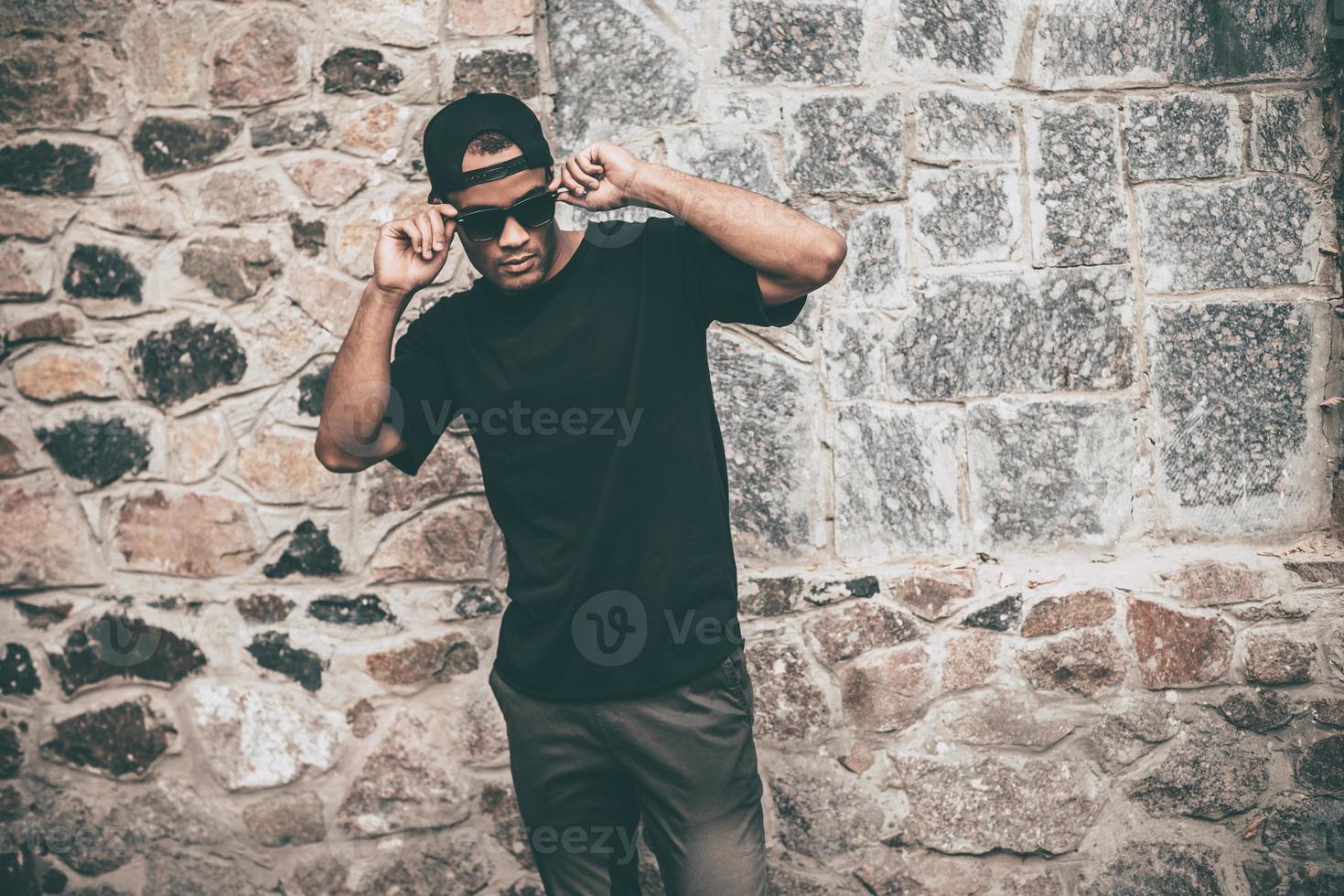 confiado en su estilo. apuesto joven africano con ropa informal ajustando sus gafas de sol mientras está de pie contra la pared de piedra al aire libre foto