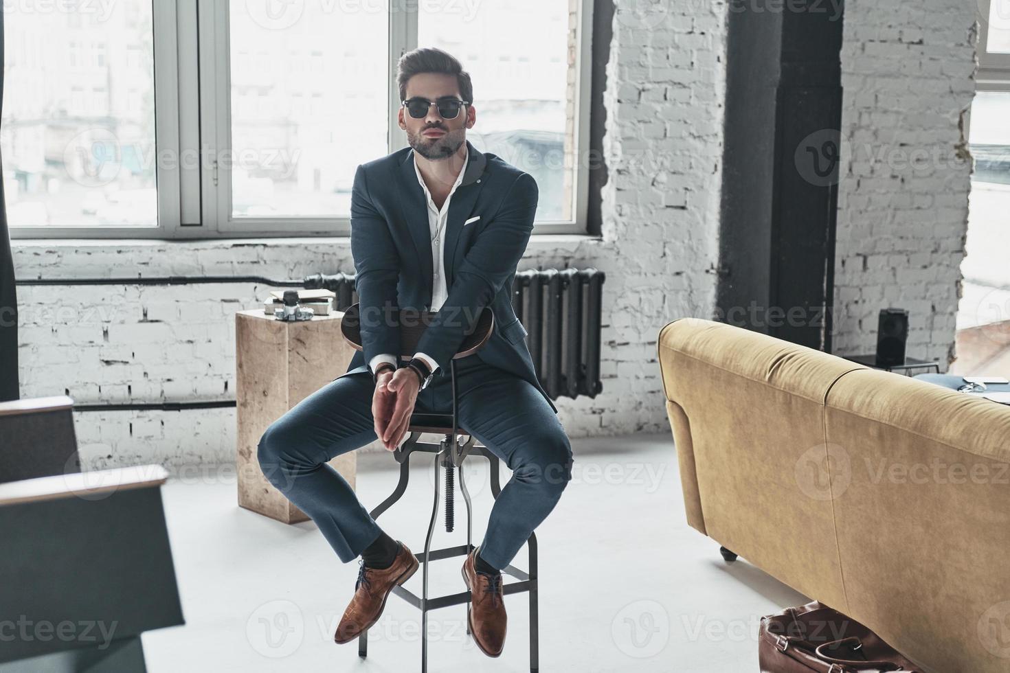 tiene estilo. apuesto joven con traje completo y anteojos manteniendo las manos juntas mientras se sienta en el taburete foto