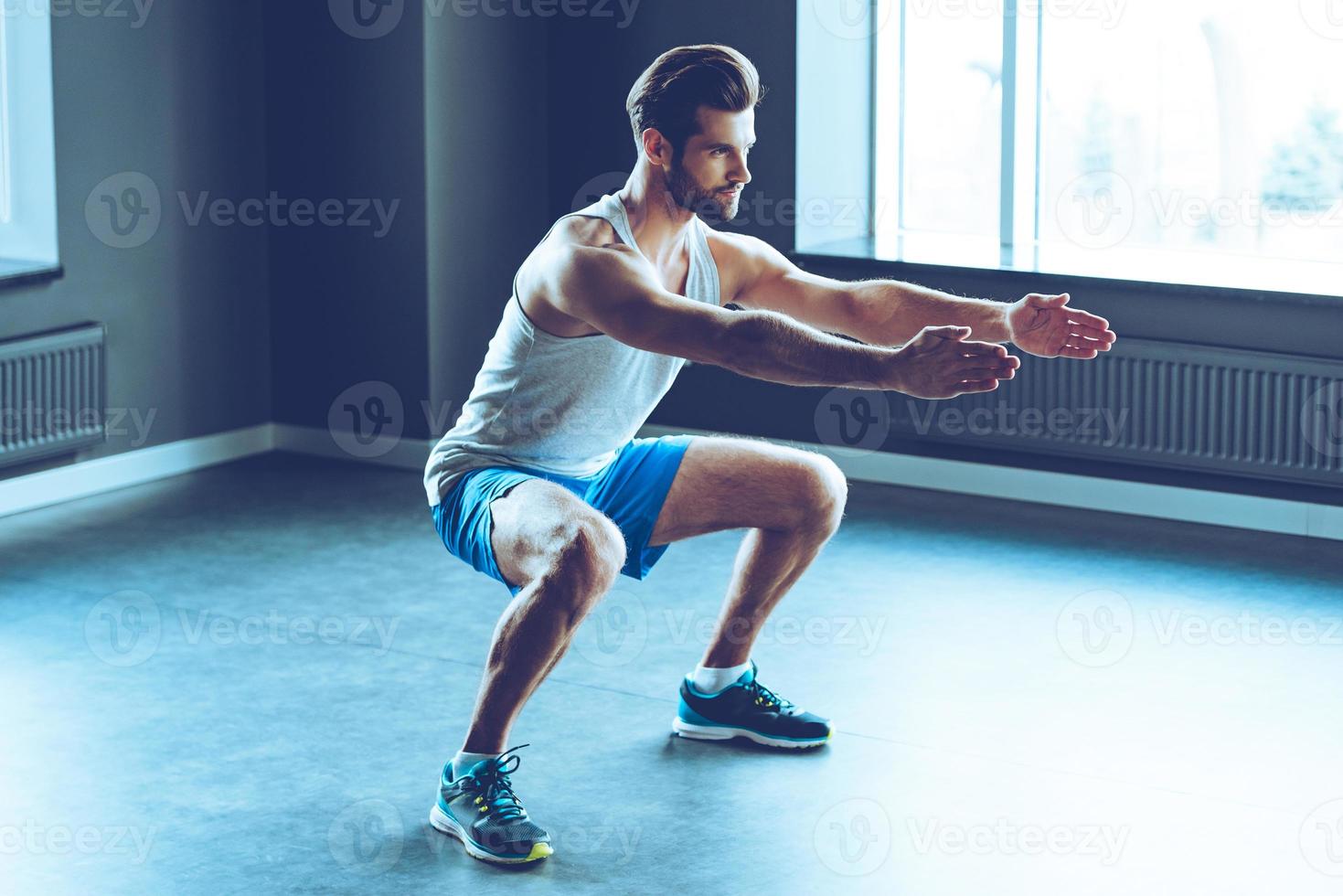 Perfect squat. Full length of young handsome man in sportswear doing squat at gym photo