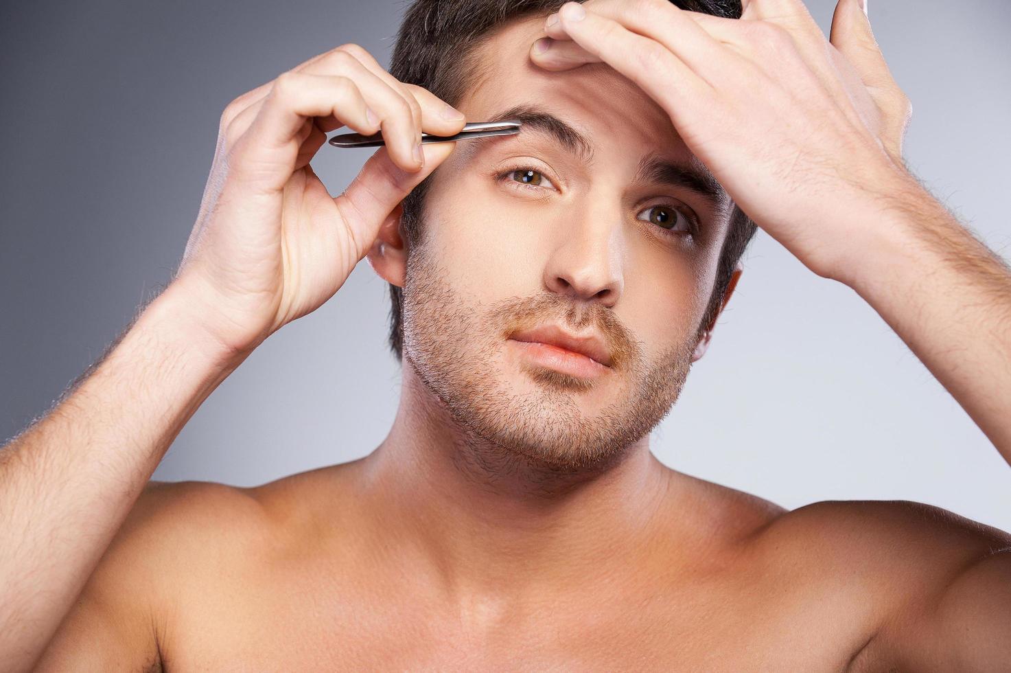 hombre depilando sus cejas. un joven apuesto que se depila las cejas y mira la cámara mientras está aislado en un fondo gris foto