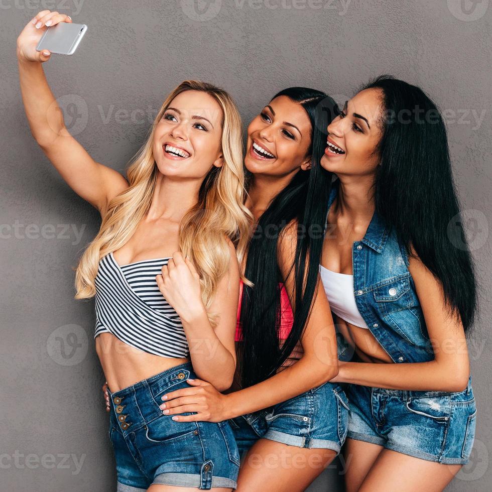Friendly selfie. Side view of three beautiful women bonding to each other and smiling while making selfie against grey background photo