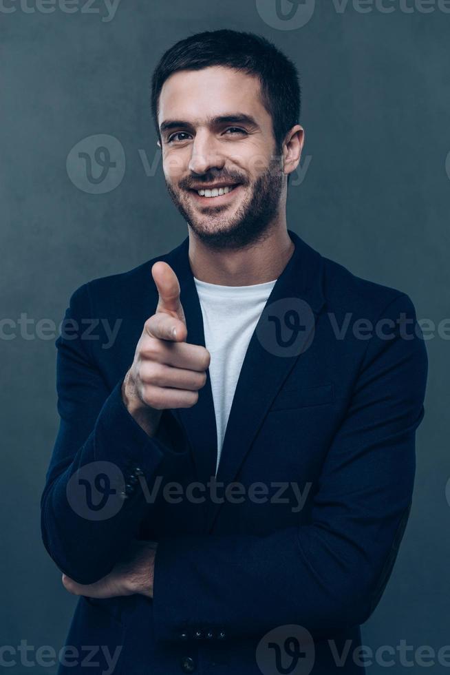 You are awesome Cheerful young man pointing you and looking at camera while standing against grey background photo