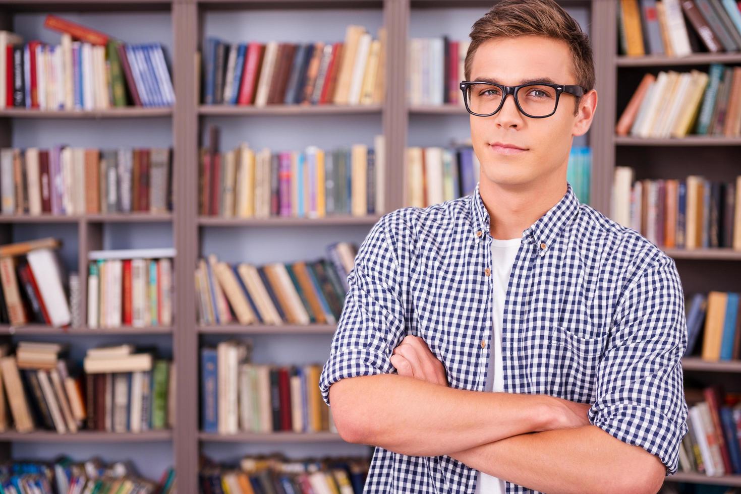 Smart and confident student. Handsome young man holding books and smiling while standing in library photo