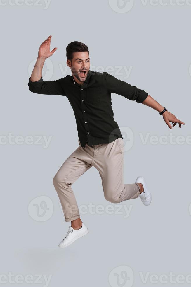 Feeling free to do anything. Full length of playful young man gesturing and smiling while jumping against grey background photo