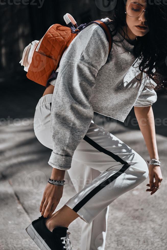 Perfect style. Beautiful young trendy woman with red backpack adjusting her shoe while standing outdoors photo