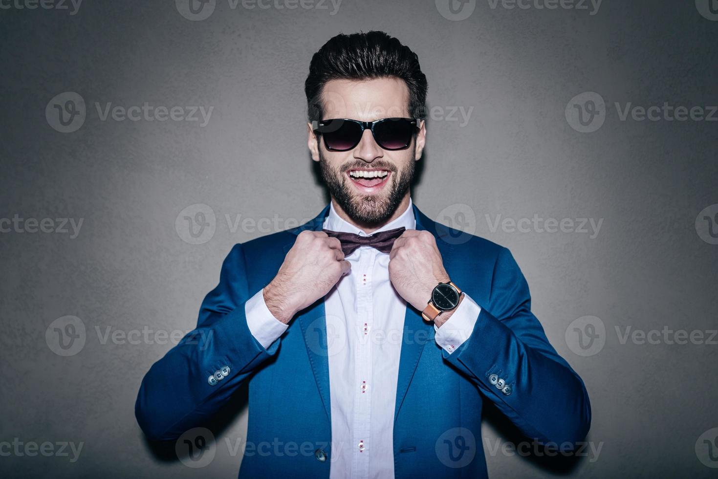 Always in good mood Close-up of handsome young cheerful man wearing sunglasses adjusting his bow tie and looking at camera with smile while standing against grey background photo