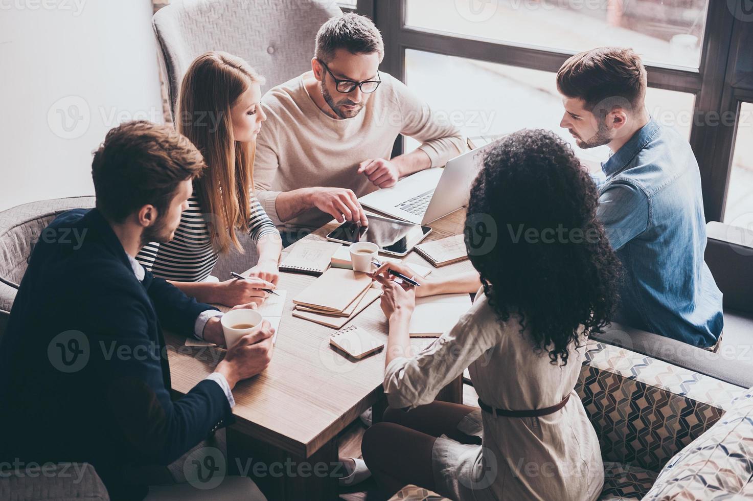 preste atención a este joven apuesto que señala una tableta digital mientras se sienta en la mesa de la oficina en una reunión de negocios con sus compañeros de trabajo foto