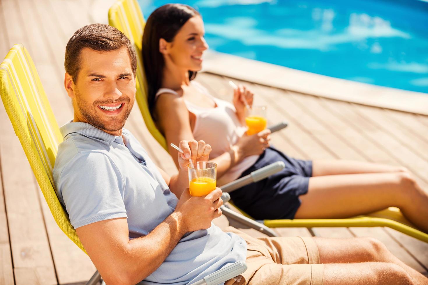 Enjoying their leisure time together. Side view of beautiful young couple sitting at the deck chairs by the pool and drinking cocktails while man looking at camera and smiling photo