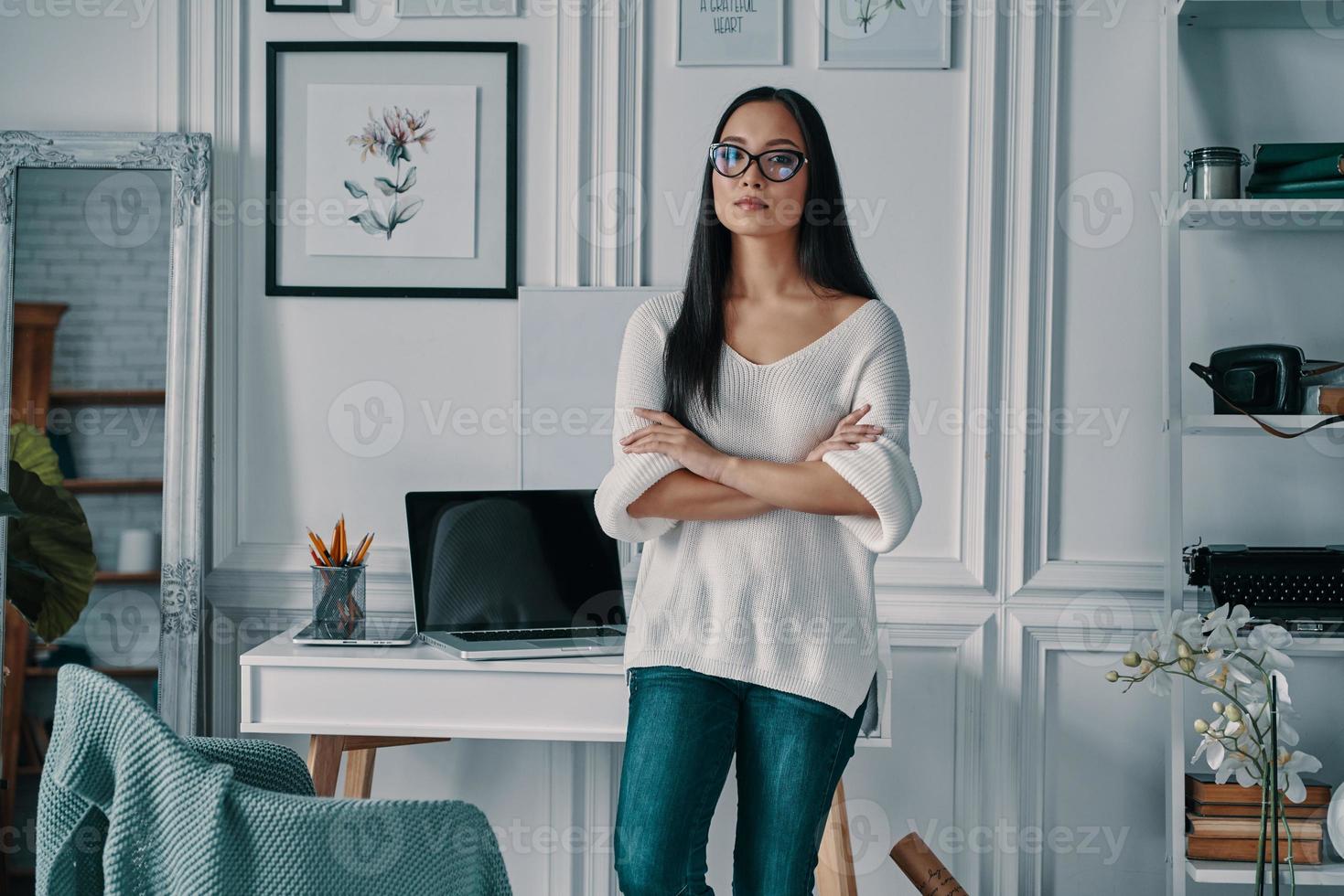 Young and full of ideas. Beautiful young woman looking at camera and keeping arms crossed while standing in home office photo