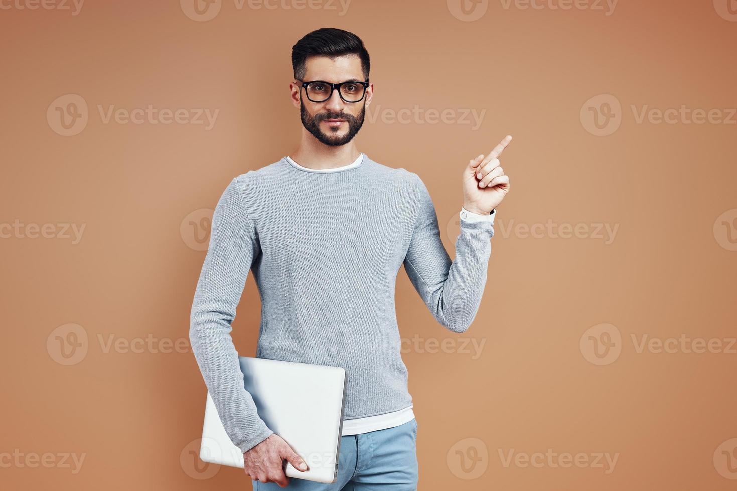 Charming young man in casual wear looking at camera and pointing copy space while standing against brown background photo