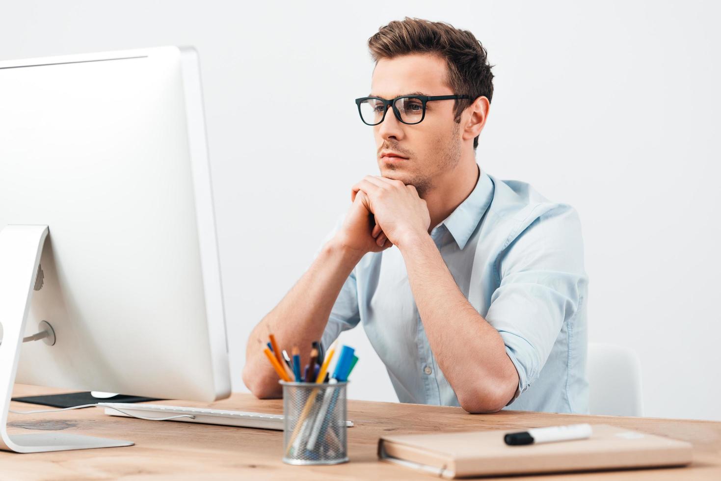 Searching for new ideas. Pensive young handsome man in glasses working on computer and keeping hand on chin while sitting at his working place photo