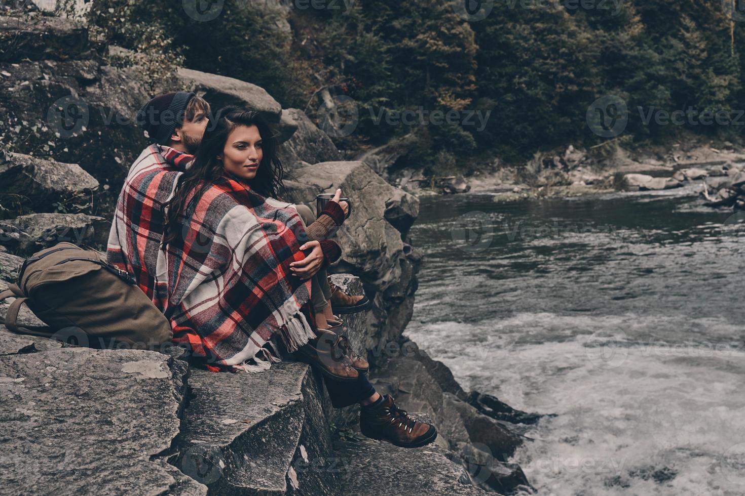 escuchando el canto de la naturaleza. hermosa pareja joven cubierta con una manta mirando hacia otro lado mientras se sienta en las rocas cerca del río foto