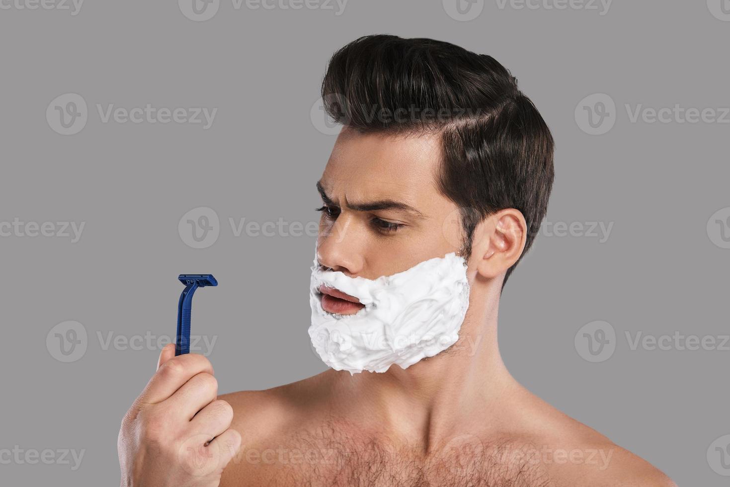Bad razor Confused young man with foam all over his face looking at razor while standing against grey background photo