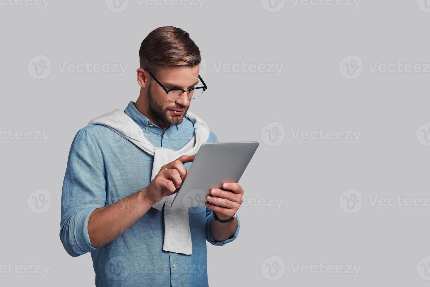 Examining his digital tablet.  Serious young man in eyeglasses working using digital tablet while standing against grey background photo