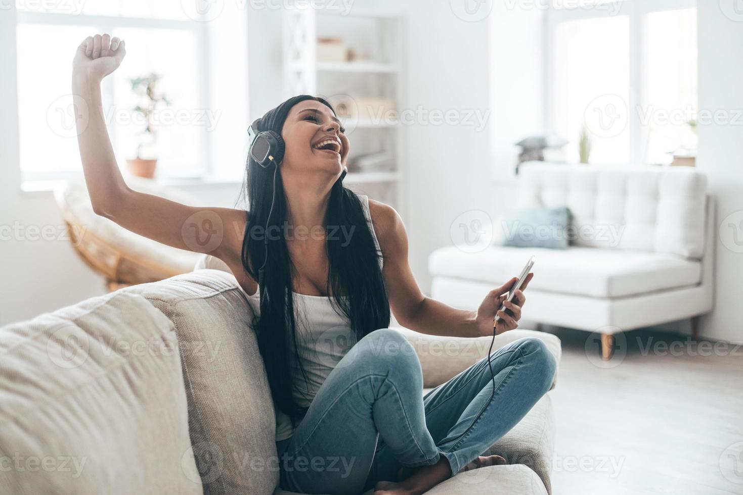 disfrutando de la música favorita mujer joven feliz en auriculares sosteniendo un teléfono inteligente y gesticulando mientras se sienta en el sofá en casa foto