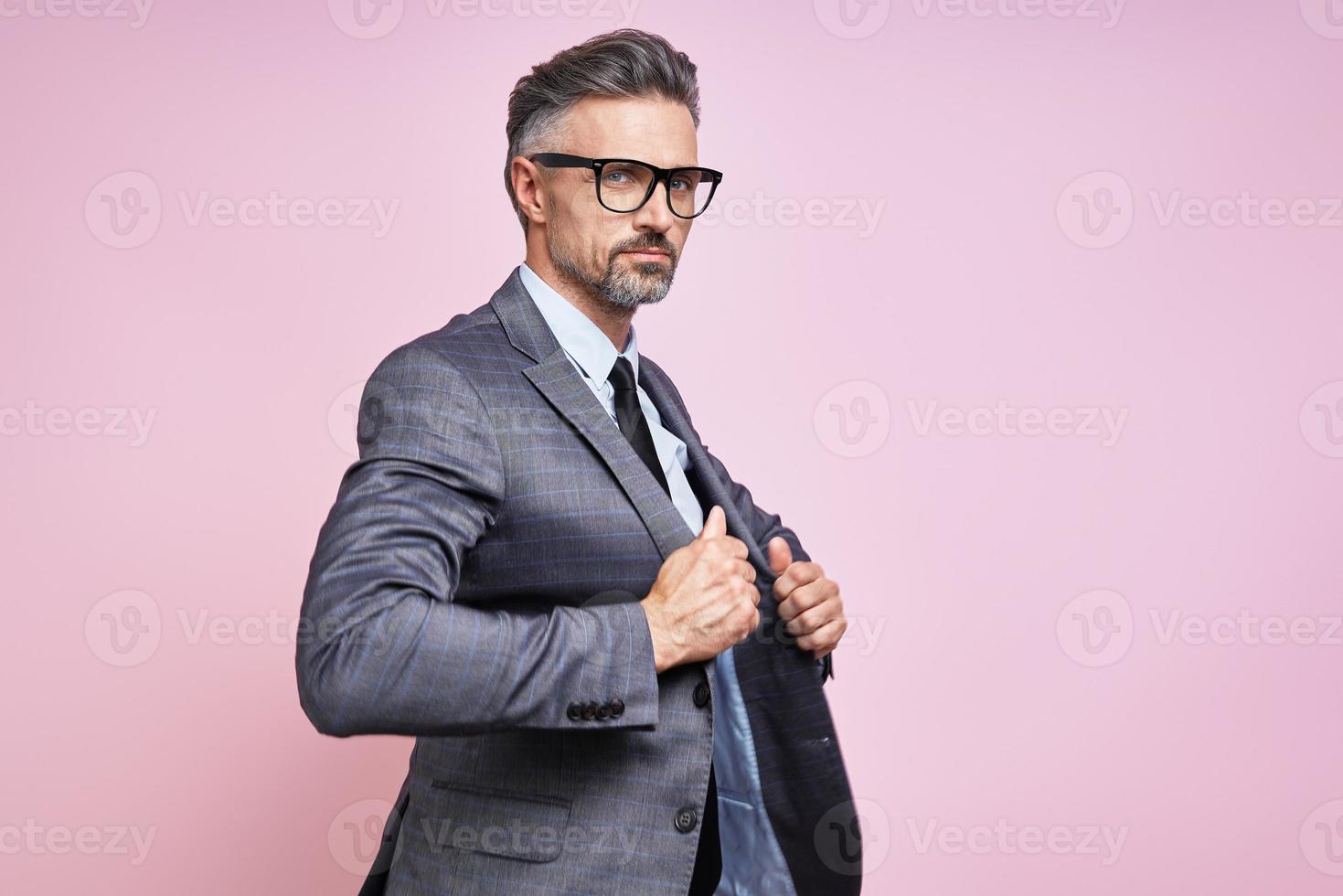 Handsome mature man in formalwear adjusting his jacket while standing against pink background photo