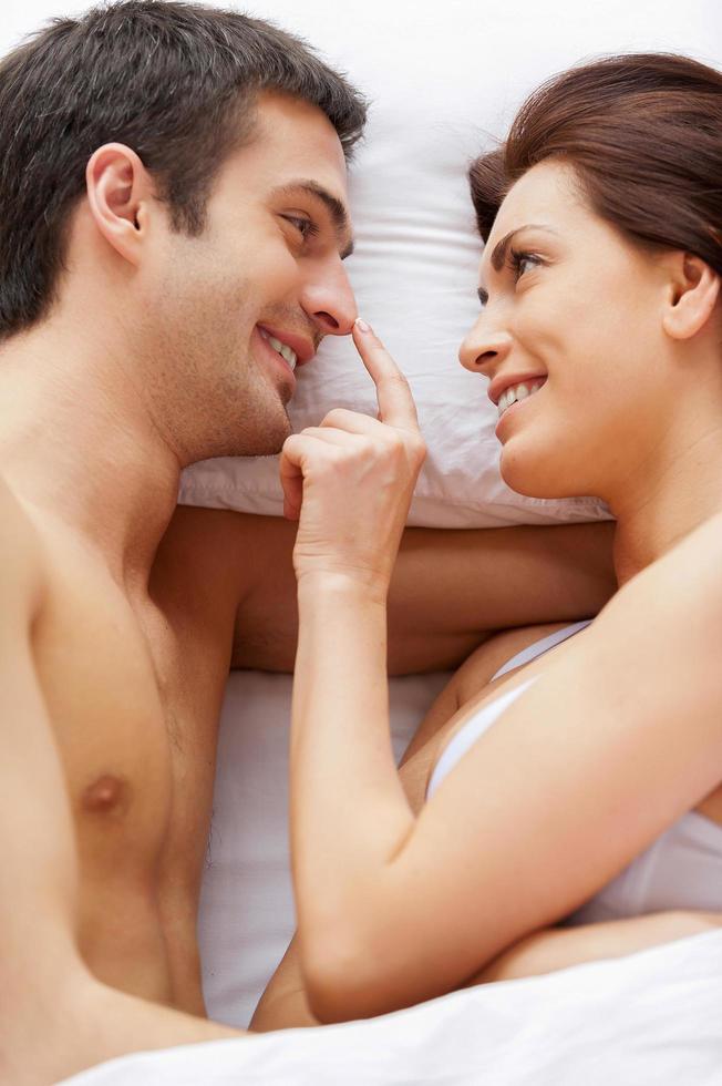 Playful couple in bed. Top view of beautiful young loving couple lying in bed while woman touching her boyfriend nose photo