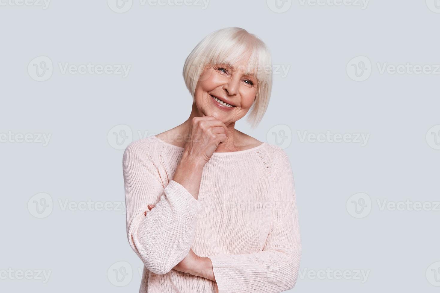 verdadera belleza femenina. hermosa anciana manteniendo la mano en la barbilla y sonriendo mientras está de pie contra el fondo gris foto