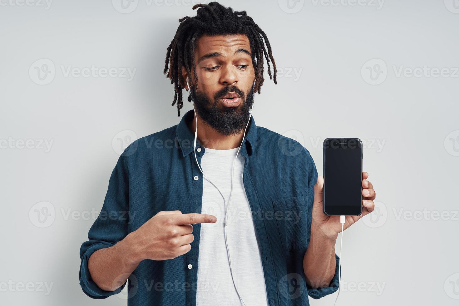 Charming young African man in casual wear pointing copy space and listening music while standing against grey background photo