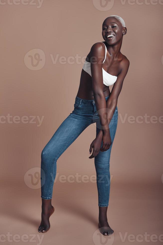 estilo africano. toda la longitud de una atractiva joven africana mirando a la cámara y sonriendo mientras se enfrenta a un fondo marrón foto