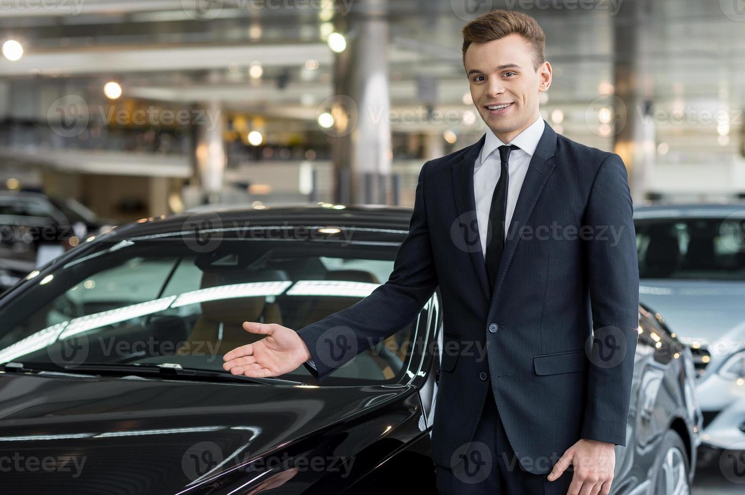 Let me show our new model. Handsome young classic car salesman standing at the dealership and pointing car photo