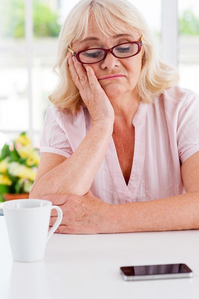 Waiting for a call. Depressed senior woman looking at her mobile phone laying on the table and leaning her head on hand photo