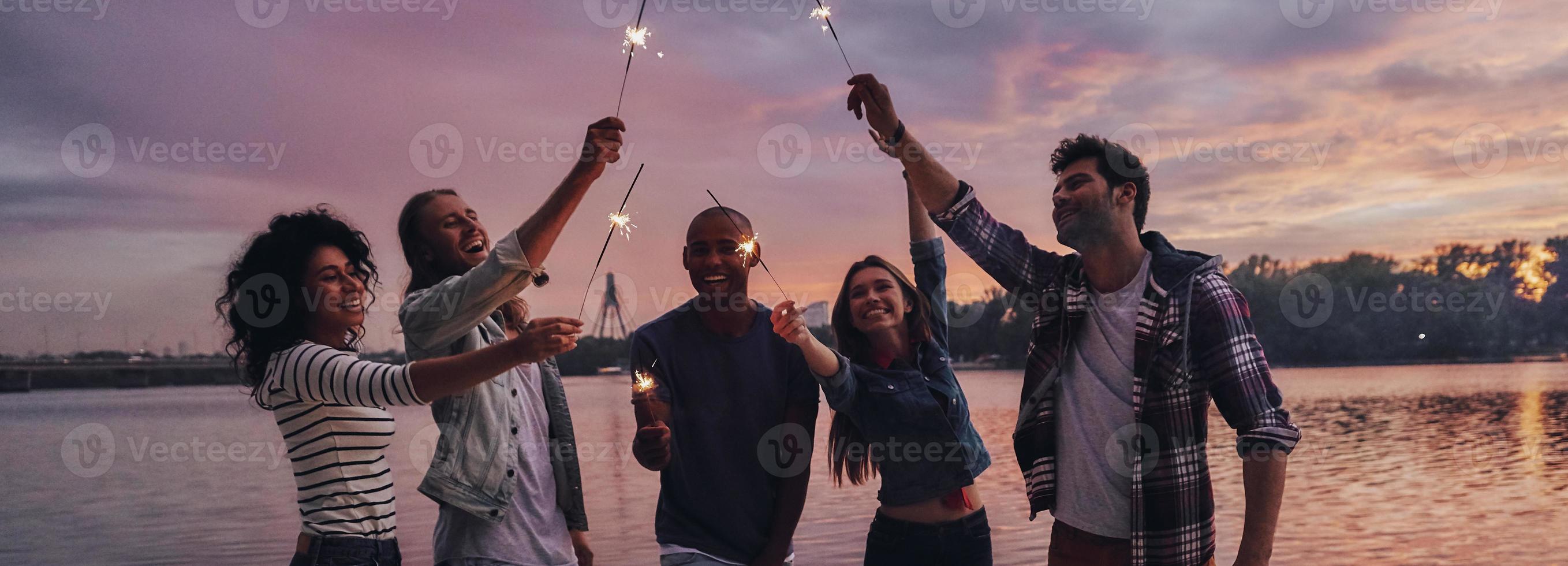 creando recuerdos felices. toda la longitud de los jóvenes en ropa casual sonriendo y sosteniendo chispas mientras están de pie en el muelle foto