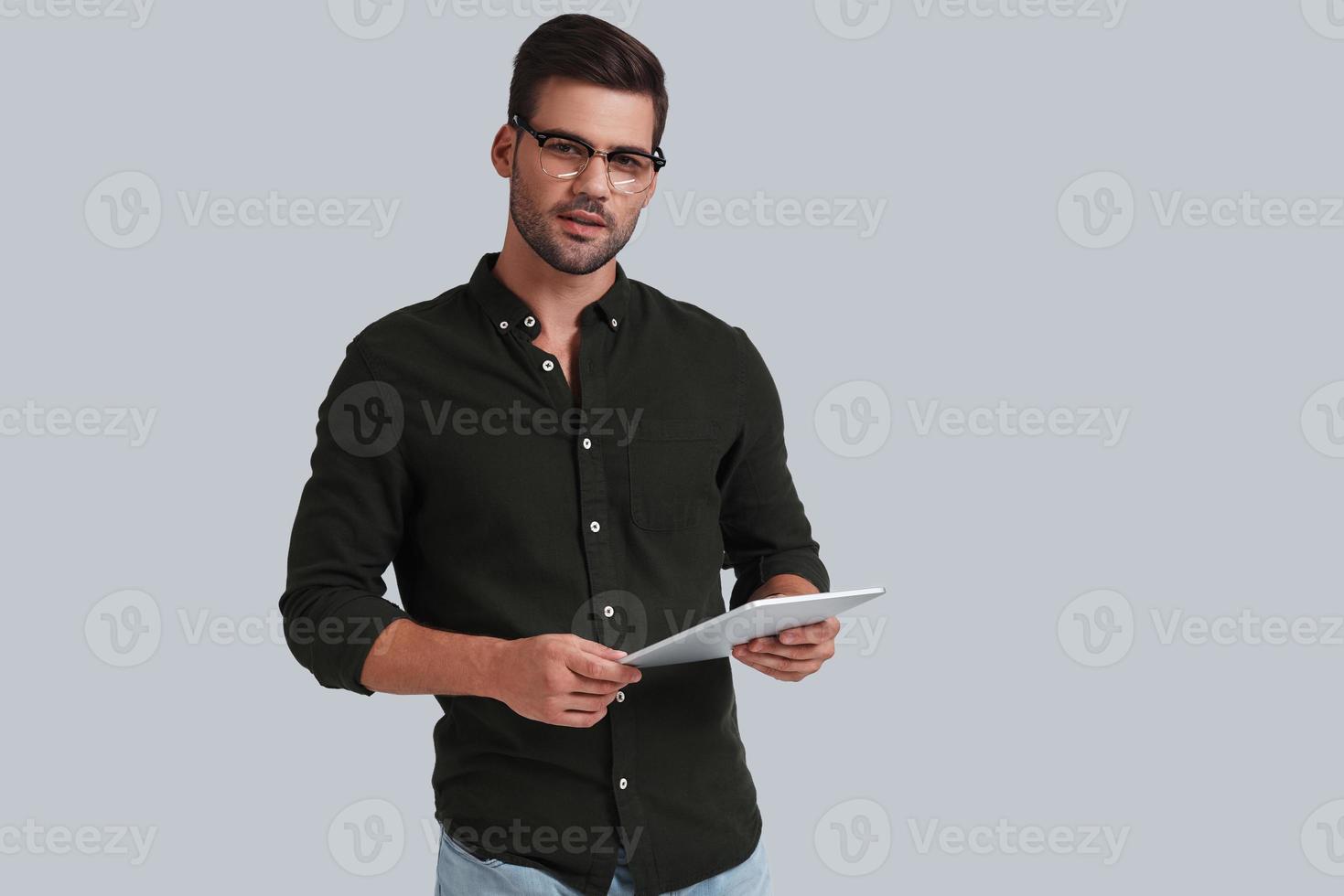 Good looking young man in eyeglasses holding his digital tablet and looking at camera while standing against grey background photo