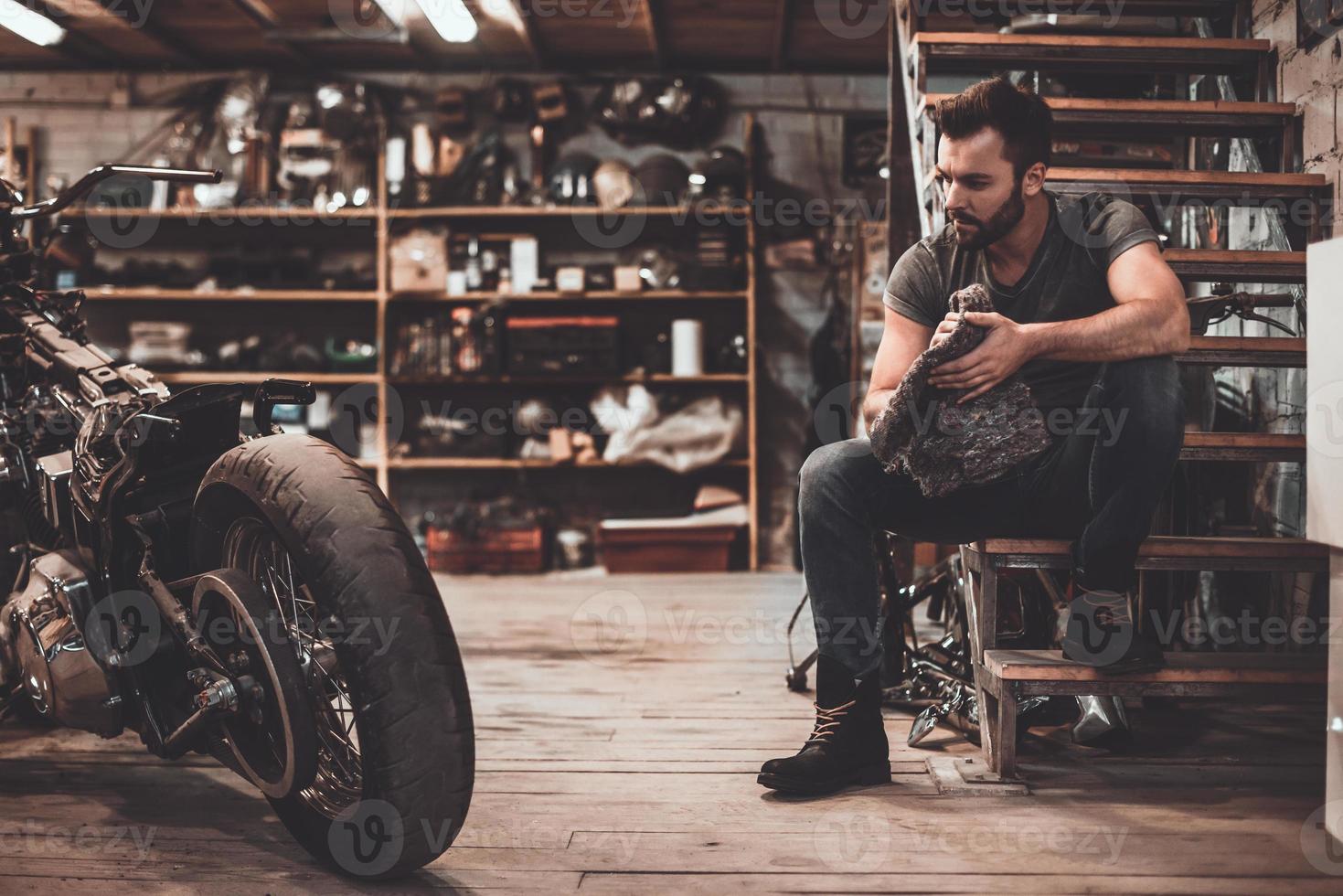 mecánico confiado. joven seguro de sí mismo sosteniendo un trapo y mirando la motocicleta mientras se sienta cerca de ella en el taller de reparación foto