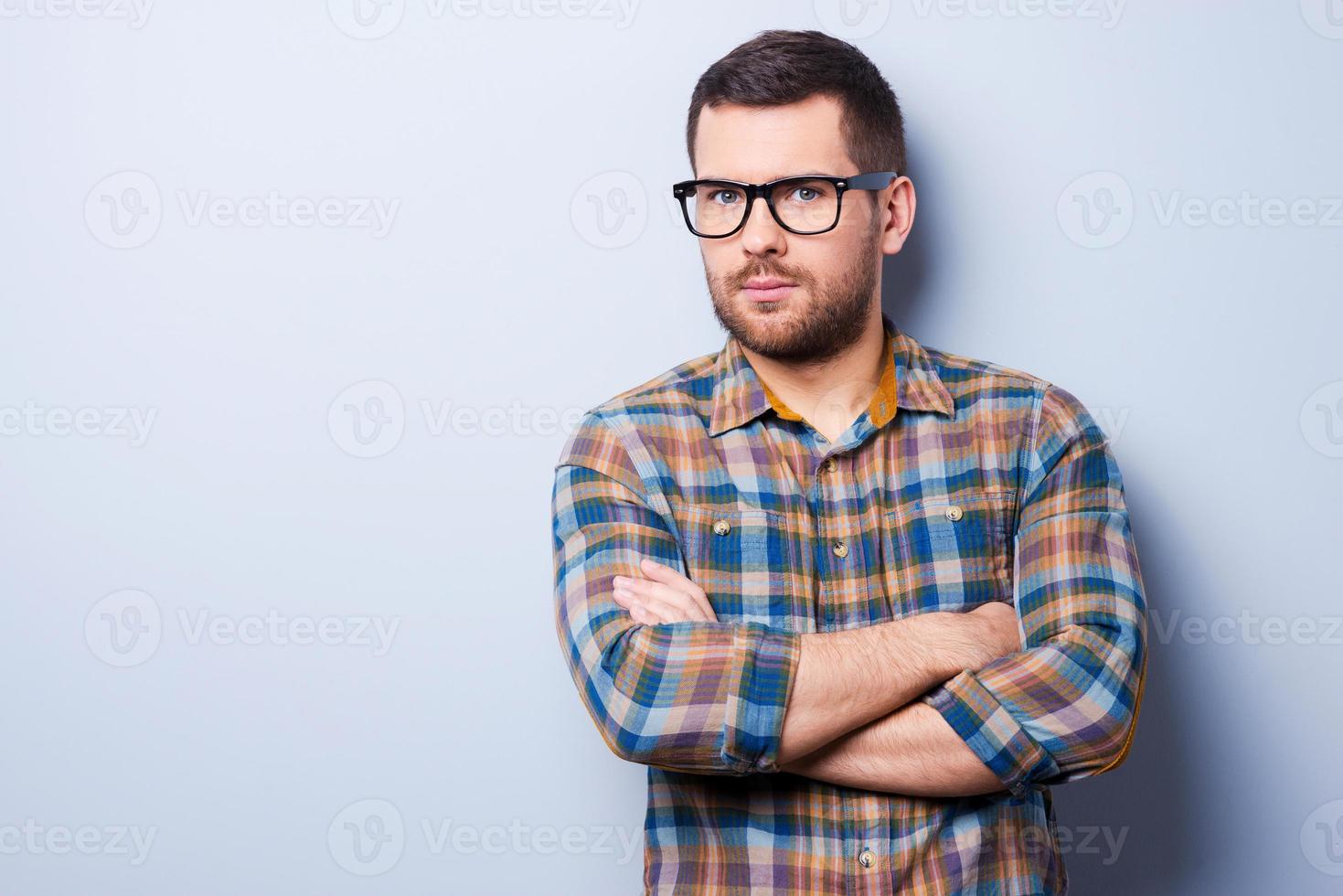 Confident and creative. Serious young man keeping arms crossed and looking at camera while standing against grey background photo