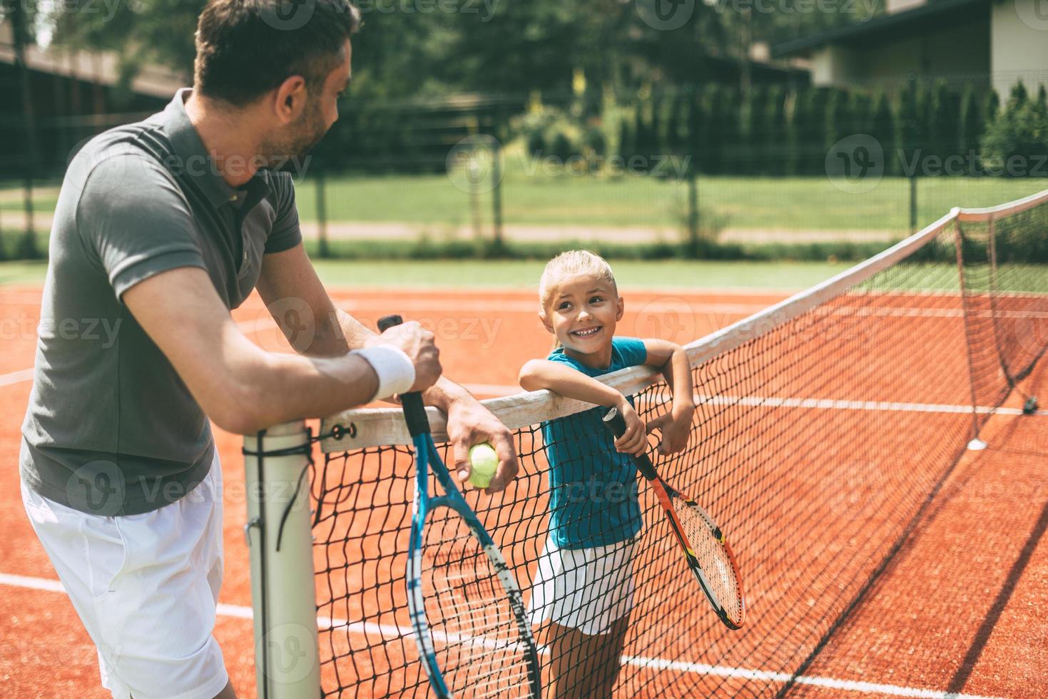 listo para jugar alegre padre e hija apoyándose en la red de tenis y mirándose con una sonrisa foto