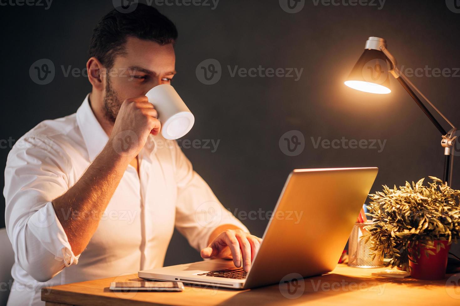 Rebooting his energy to work more. Confident young man drinking coffee while sitting at his working place at night time photo