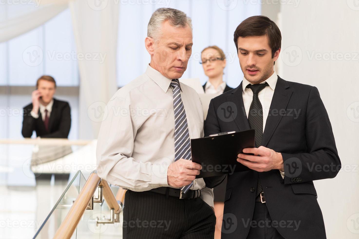Asking an advice. Two men in formalwear discussing something while moving by staircase with people on background photo