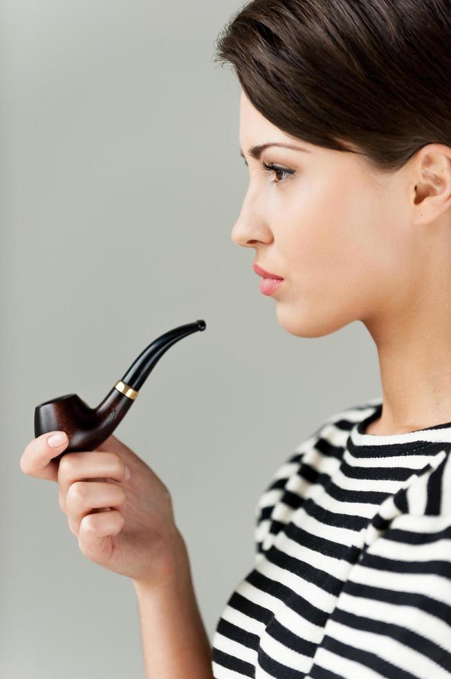 fumando. vista lateral de una atractiva mujer joven de pelo corto con ropa a rayas sosteniendo una pipa y mirando hacia otro lado foto