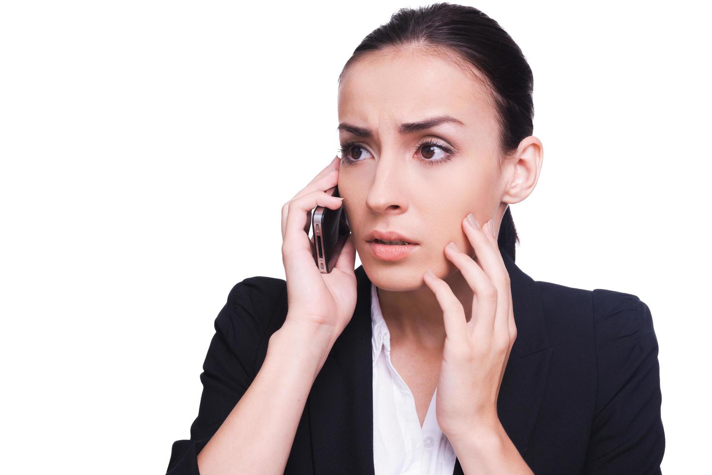 Bad news. Shocked young woman in formalwear talking on the mobile phone and touching her face with hand while standing isolated on white photo