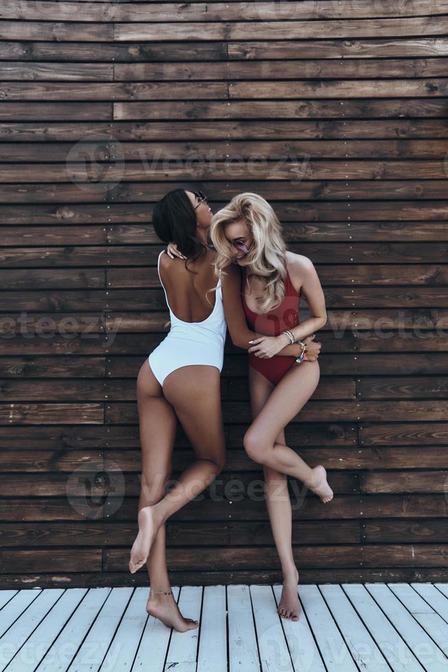 Young and full of energy. Full length of two attractive young women in swimwear laughing while standing against the wooden wall outdoors photo