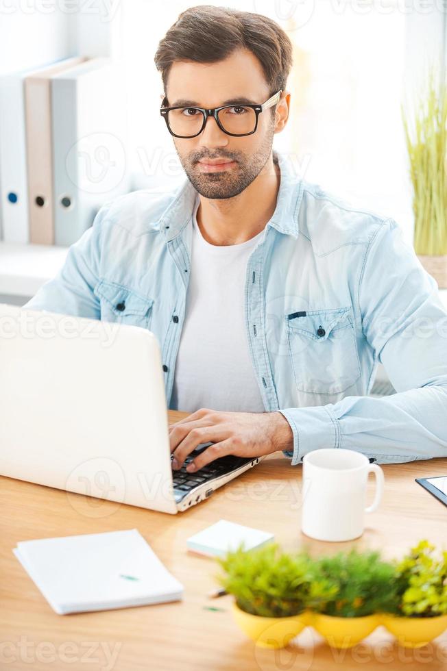 trabajando en un proyecto creativo. un joven apuesto con camisa y anteojos trabajando en una laptop mientras se sienta en su lugar de trabajo foto