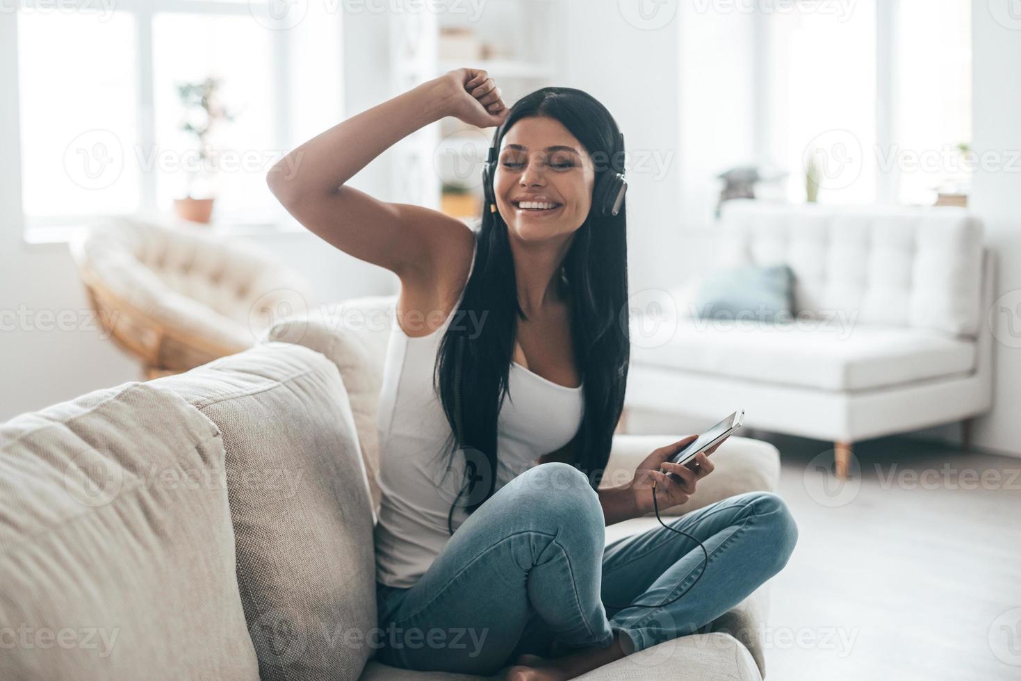Enjoying best song ever Attractive young woman in headphones holding smart phone and gesturing while sitting on the couch at home photo