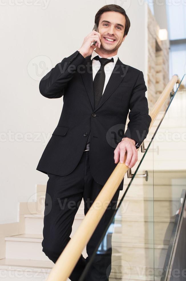 Always in touch. Cheerful young man in formalwear talking on the mobile phone while moving down by staircase photo