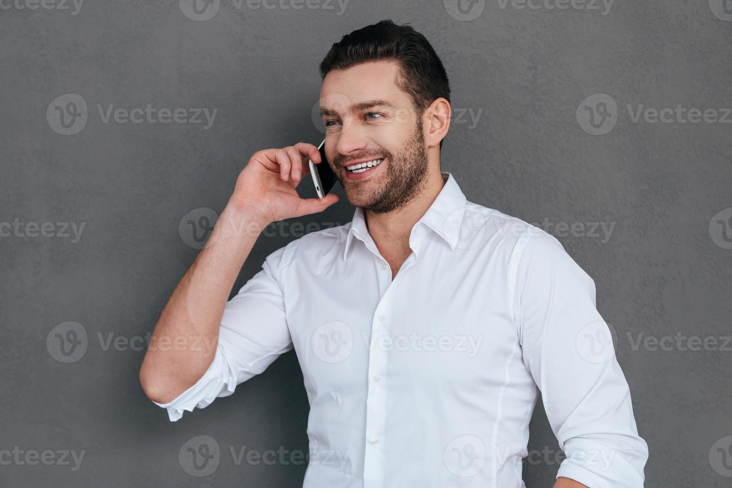 Good business talk. Handsome young man talking on the mobile phone and smiling while standing against grey background photo
