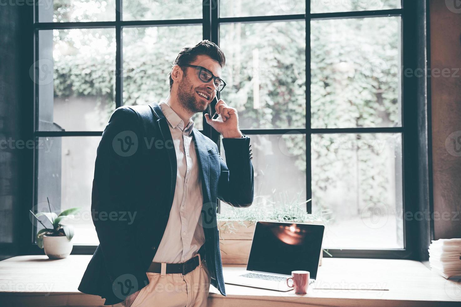Always in touch with colleagues. Confident young man talking on the mobile phone and smiling while standing in front of the big window in office or cafe photo