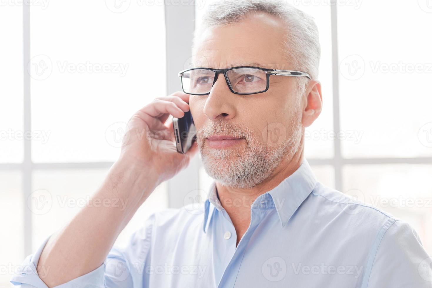 Tell me good news Serious grey hair senior man in shirt talking on the mobile phone and looking away while standing in front of the window photo
